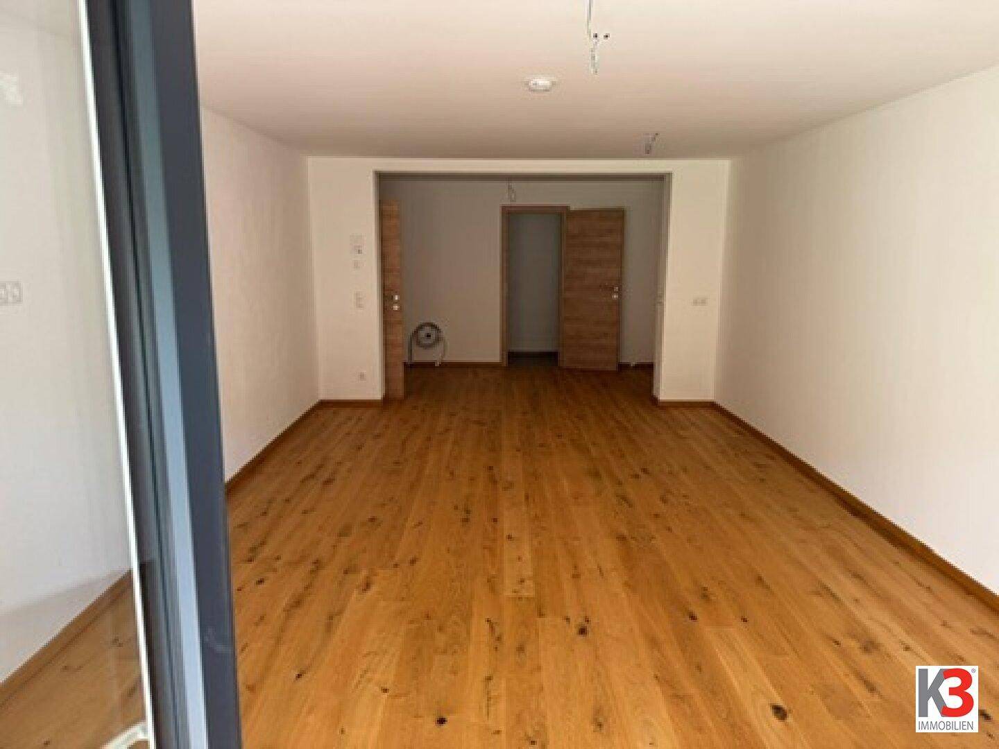 Heller, leerer Raum mit neuem Holzboden und Zugang zu weiteren Zimmern.