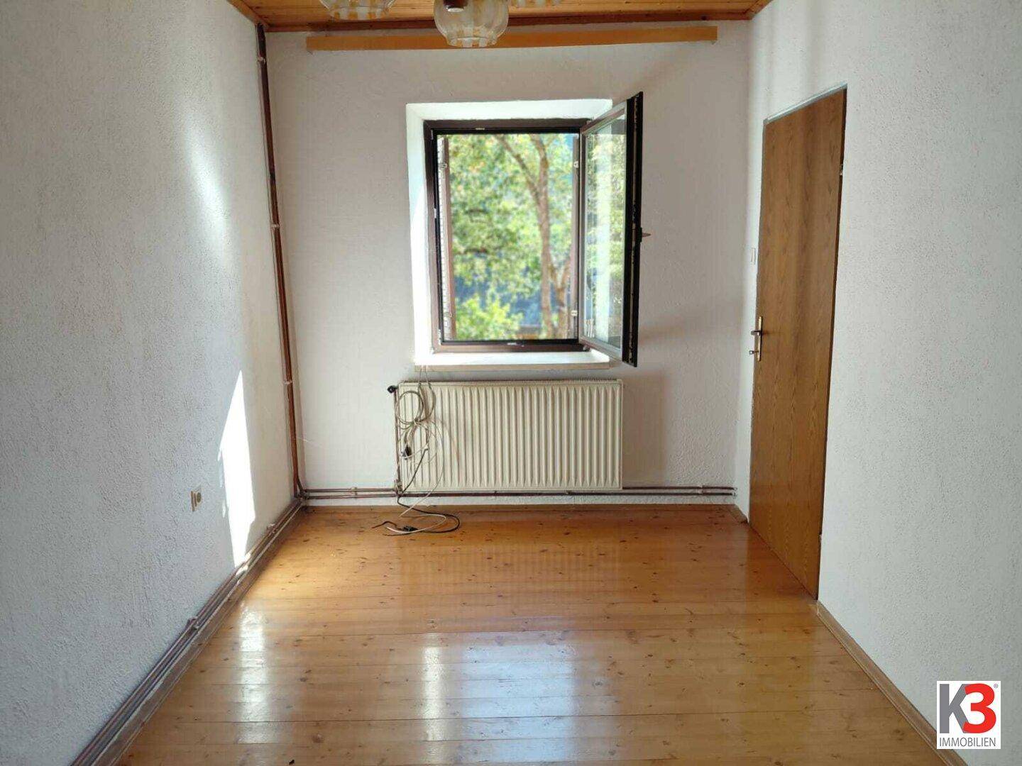 Heller Raum mit Holzboden, einem Fenster mit Blick ins Grüne und Heizkörper.