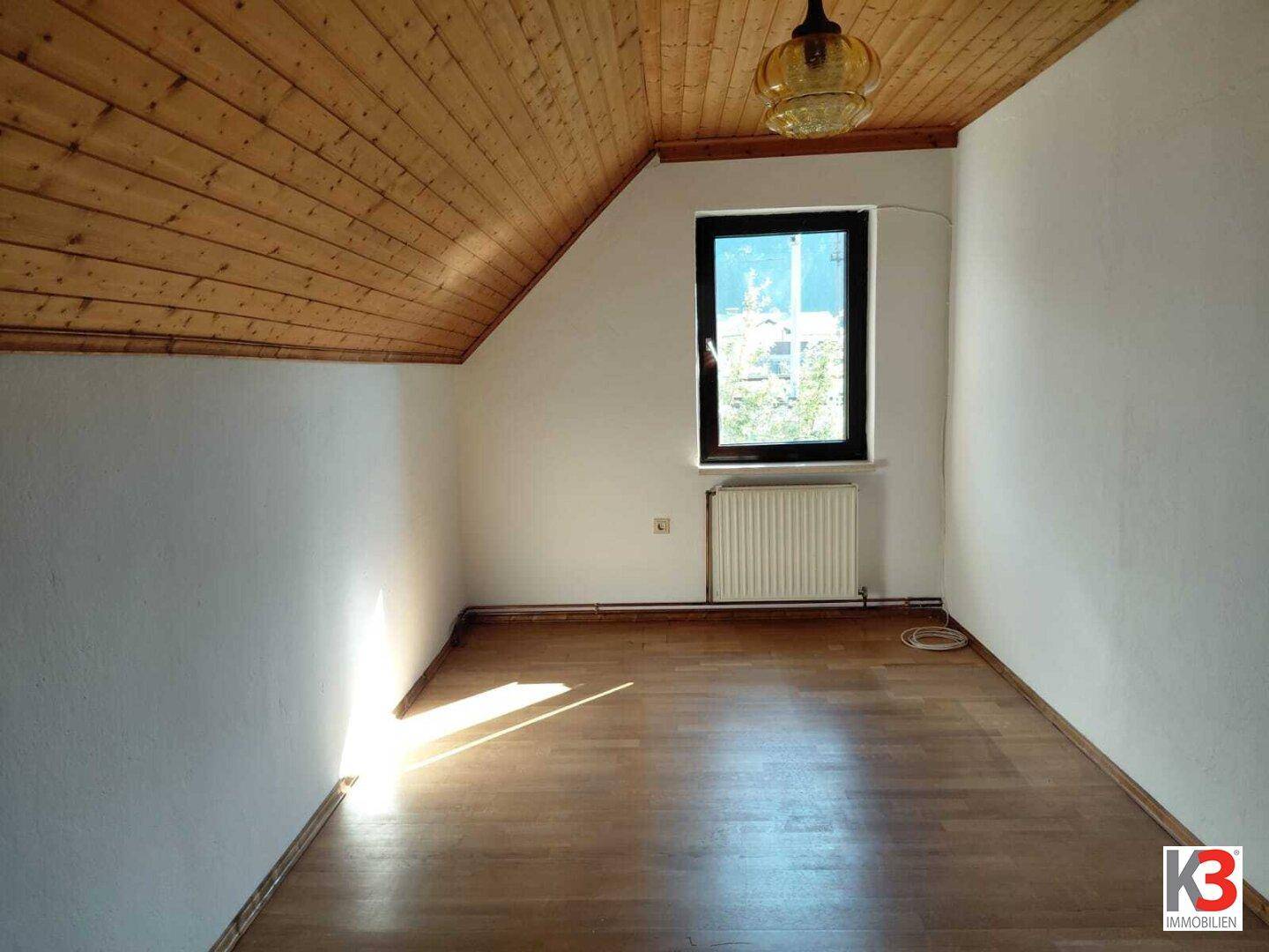 Dachgeschosszimmer mit Holzdecke, Holzboden und einem Fenster, das viel Licht hereinlässt.