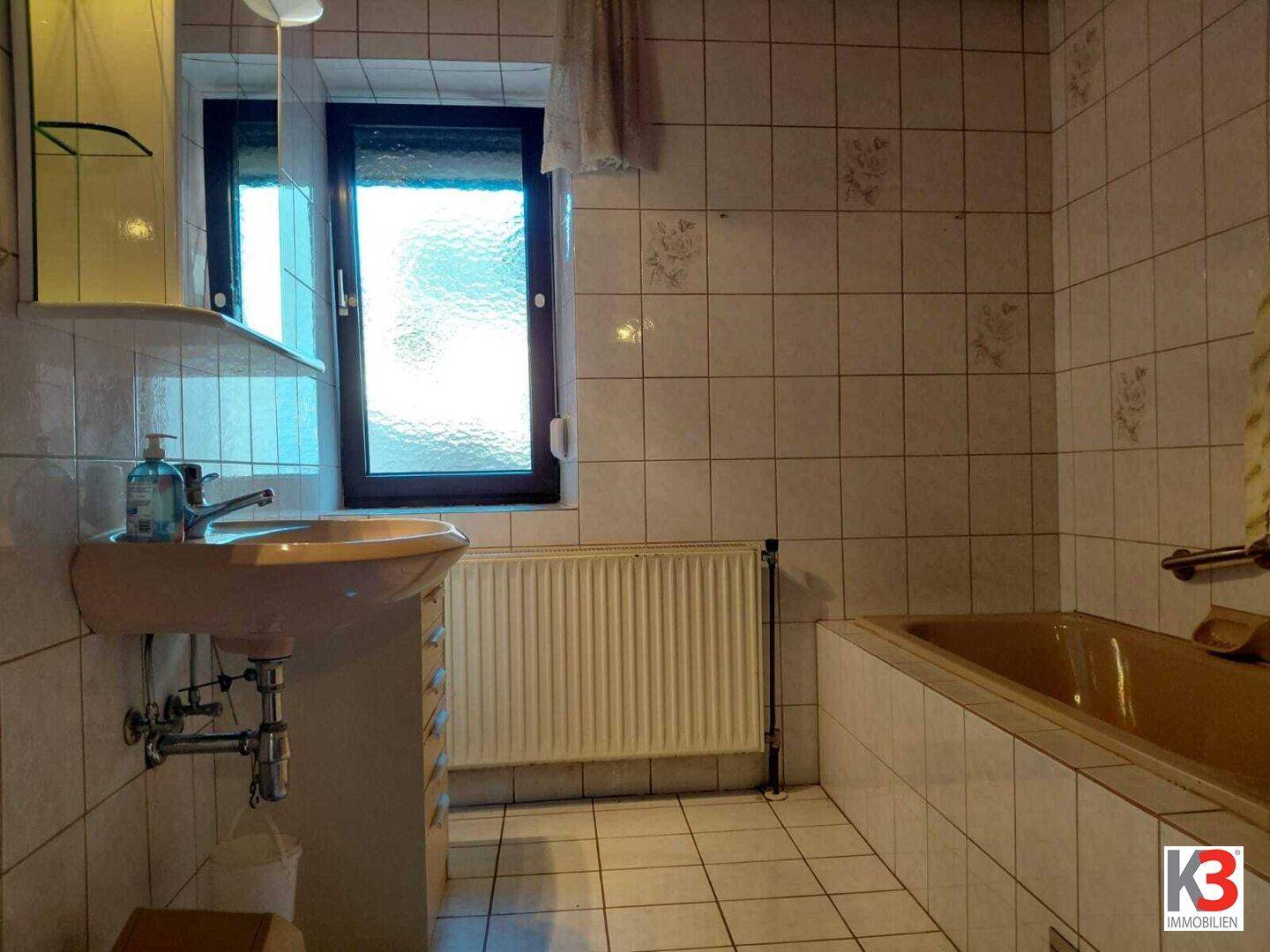 Badezimmer mit beigen Fliesen, Waschbecken, Badewanne und einem Fenster für Tageslicht.