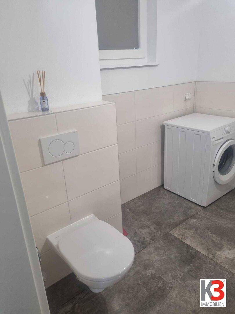 Badezimmer mit wandhängendem WC, Fenster und Waschmaschine, hell gefliest.