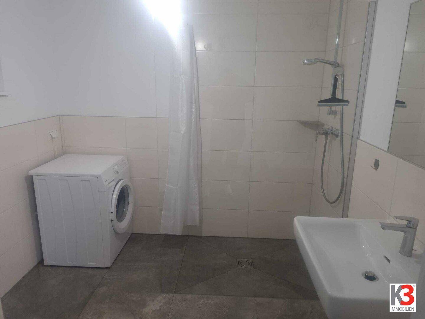 Funktionales Badezimmer mit Dusche, Waschbecken und Waschmaschine, hell gefliest.