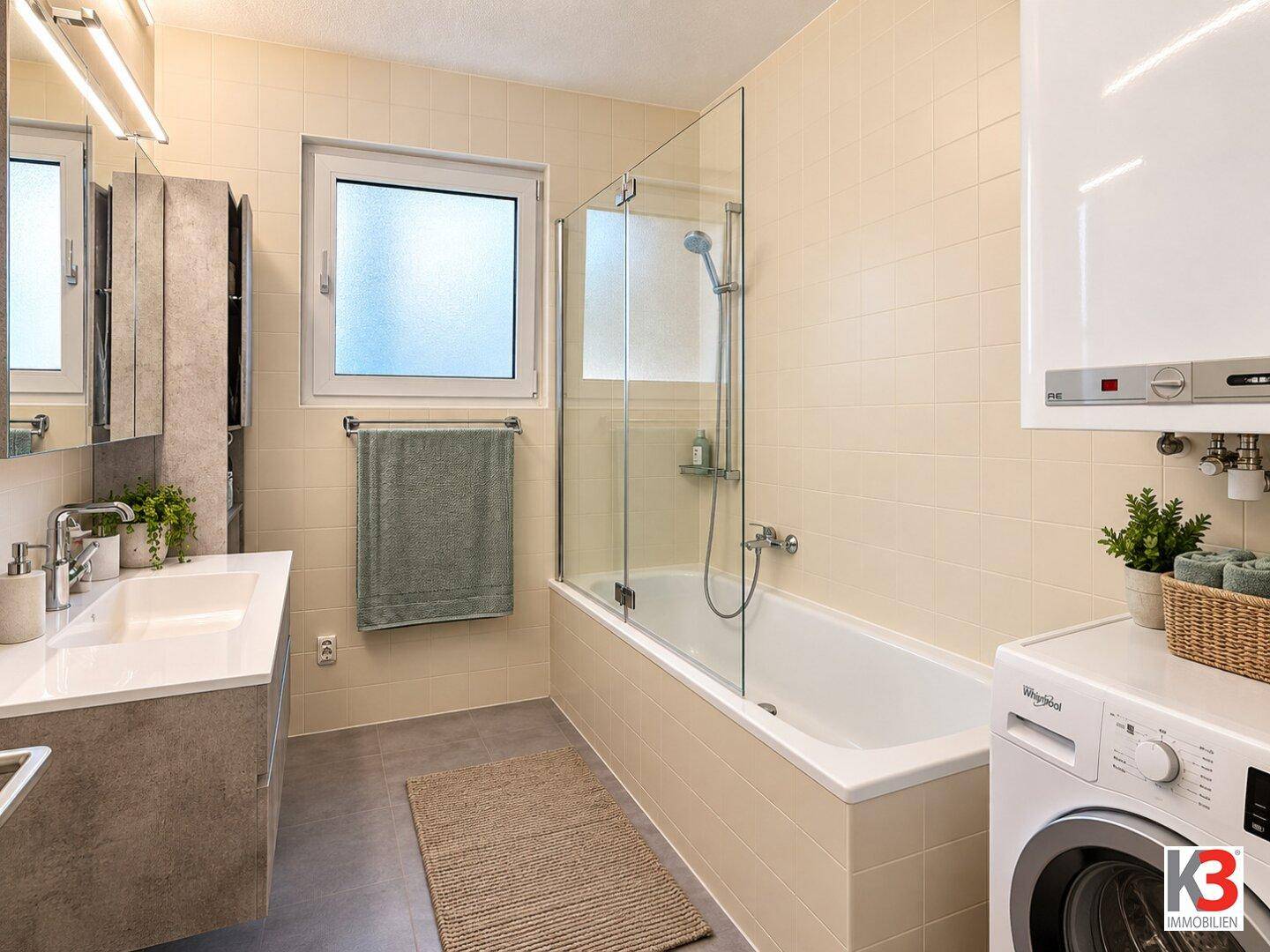 Helles Badezimmer mit Badewanne, Duschabtrennung, Einzelwaschbecken und Waschmaschine, mit Fenster.