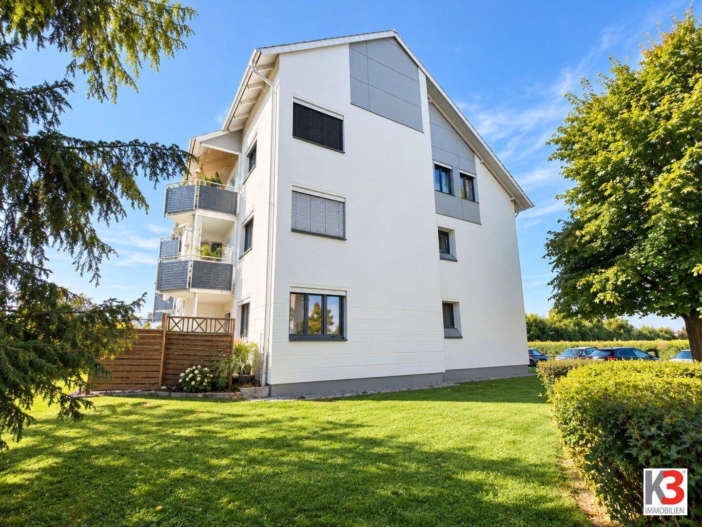 Seitenansicht des modernen Wohngebäudes mit weißer Fassade, grauen Akzenten und Balkonen, umgeben von Grünflächen.