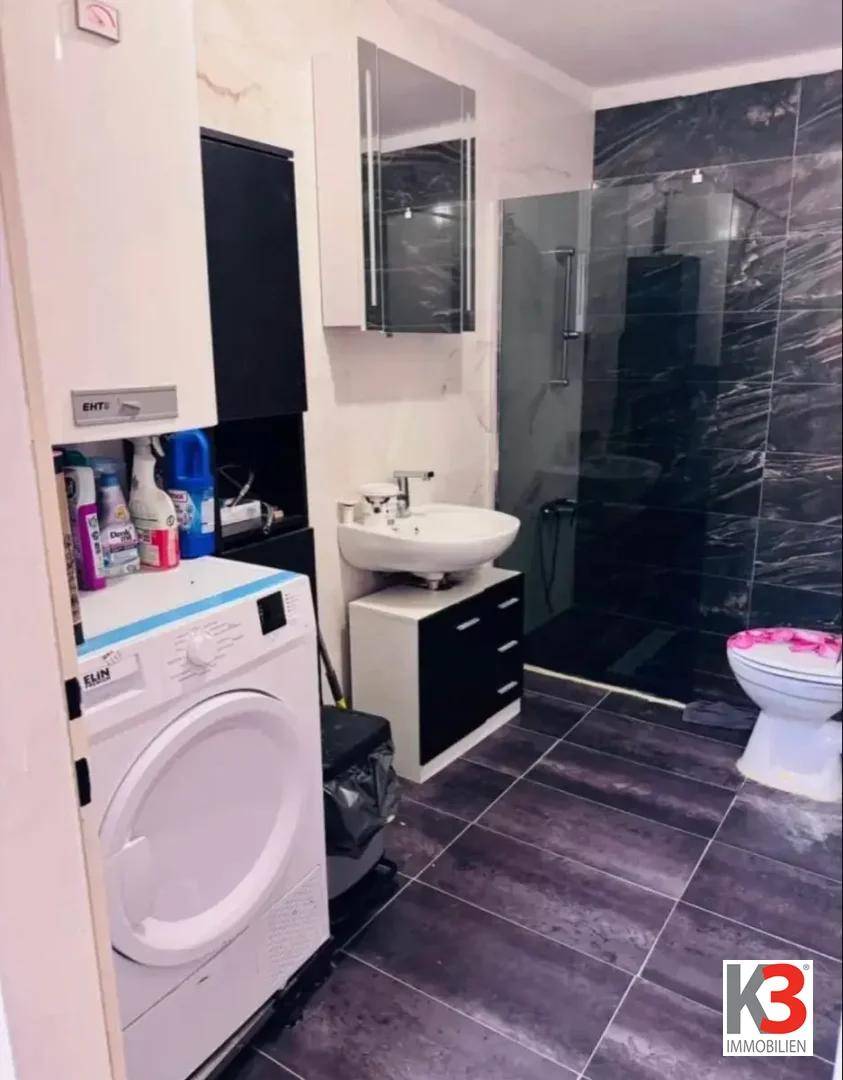 Modernes Badezimmer mit dunklen Fliesen, Duschkabine, Waschbecken und Waschmaschine.