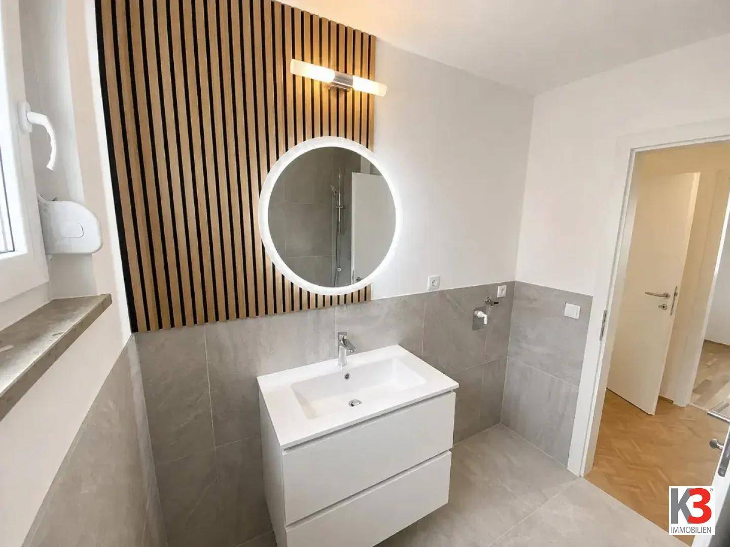 Modernes Badezimmer mit Waschtisch, beleuchtetem Rundspiegel und dekorativer Holzlamellenwand.