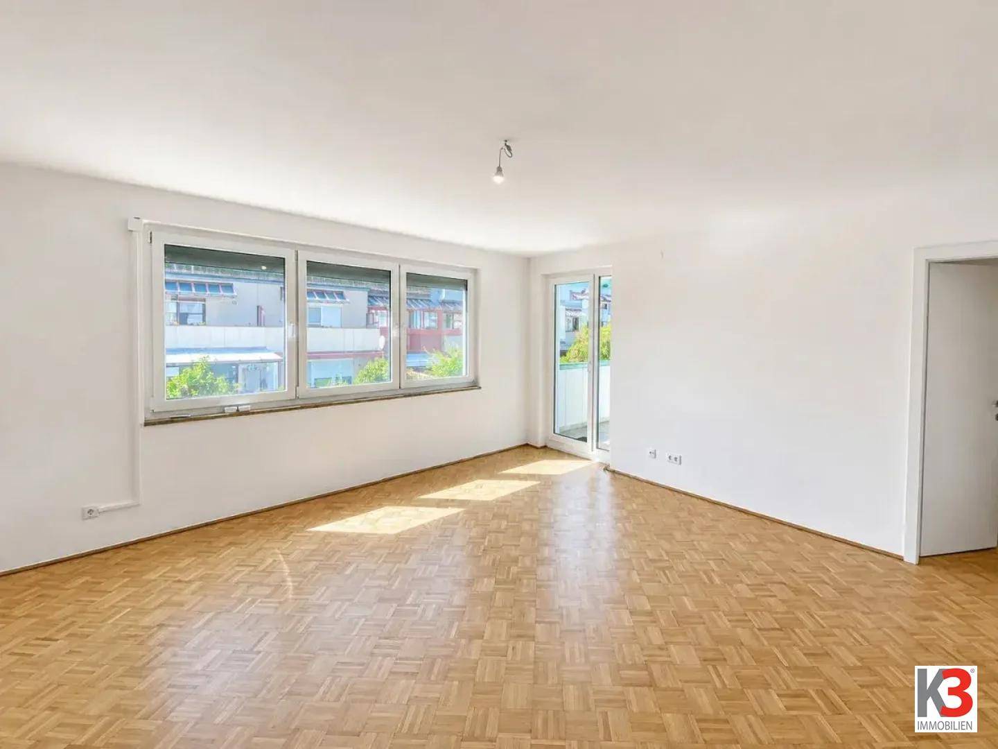 Geräumiges Wohnzimmer mit Parkettboden, großen Fenstern und Zugang zu einem Balkon oder einer Terrasse.