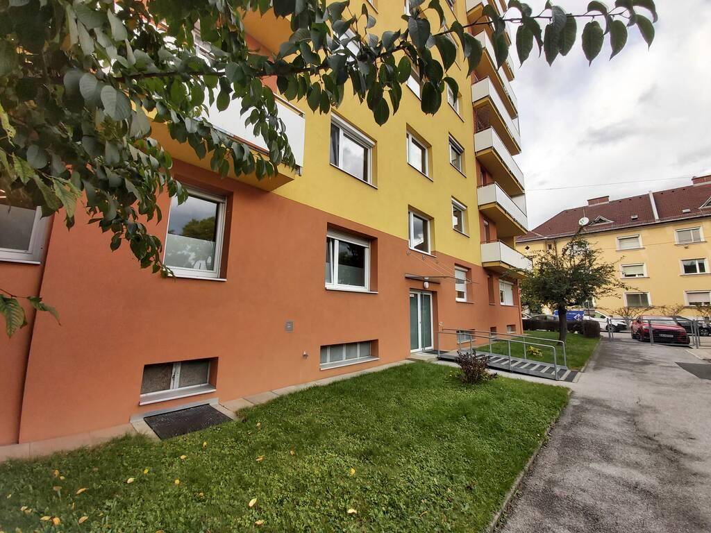 Eingangsbereich des Gebäudes mit orangefarbenem Sockel, einer Rampe für Barrierefreiheit und grüner Rasenfläche.