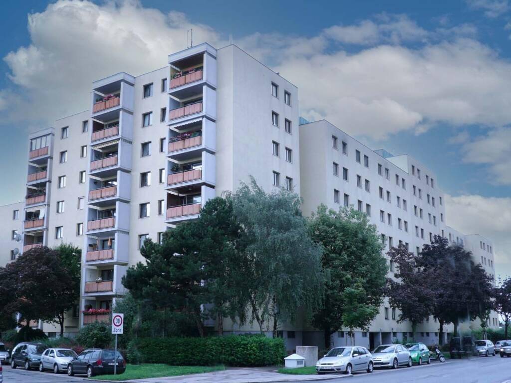 Mehrstöckiges Wohngebäude mit zahlreichen Balkonen und Bäumen an der Straße unter bewölktem Himmel.