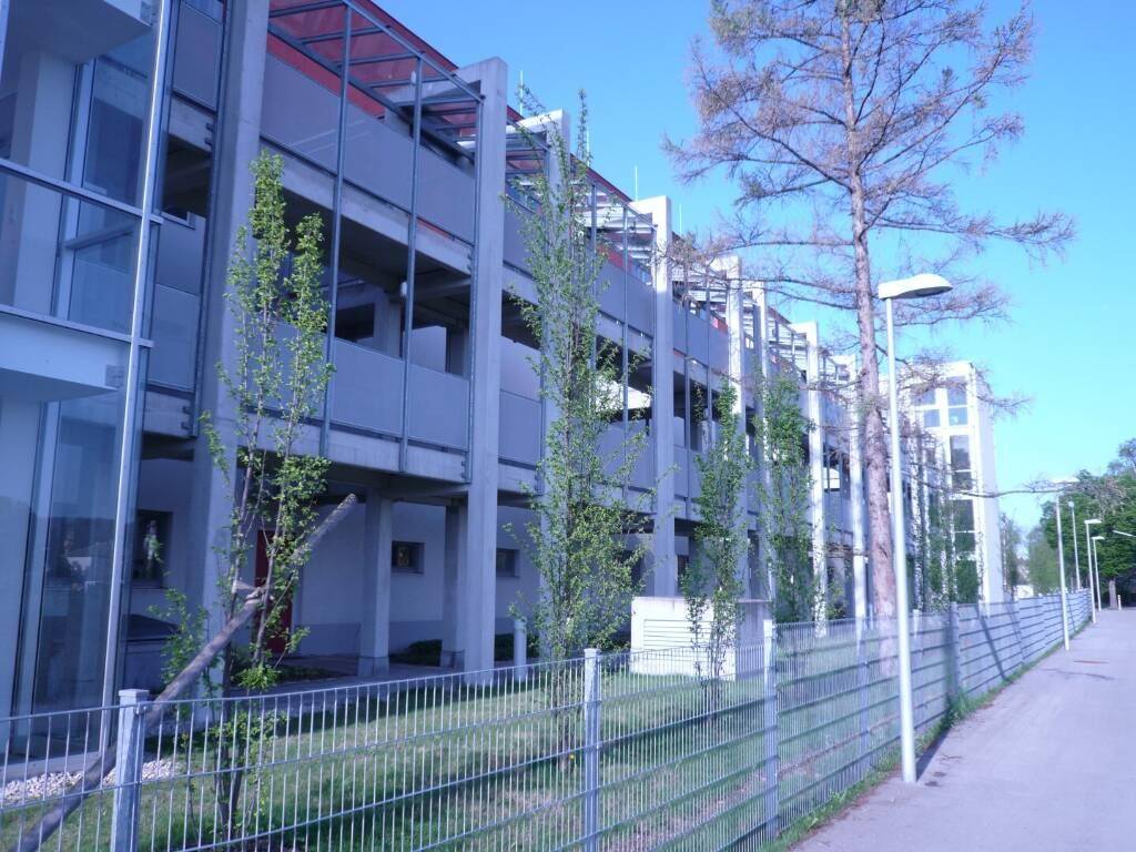 Lange Fassade eines modernen Gebäudes mit mehreren Etagen und junger Bepflanzung.