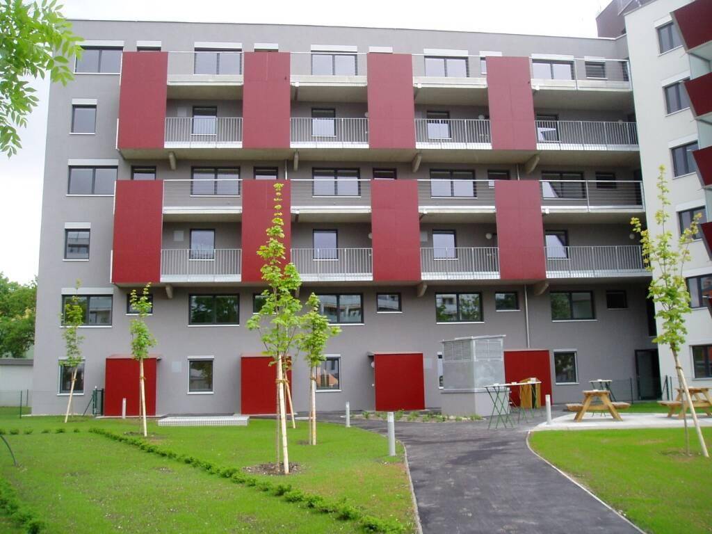 Moderne Wohnfassade mit grauen Wänden, roten Akzenten und Balkonen sowie einem gepflegten Rasenbereich.