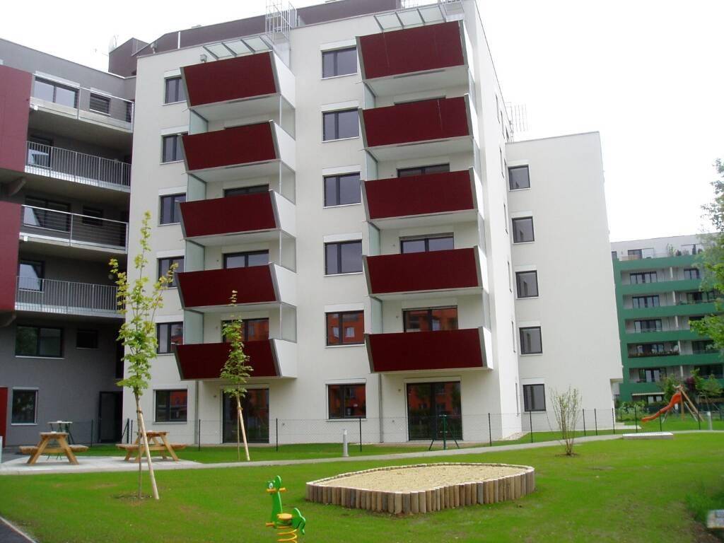 Helle Wohnfassade mit roten Balkonen und einem Spielplatz im grünen Gartenbereich.