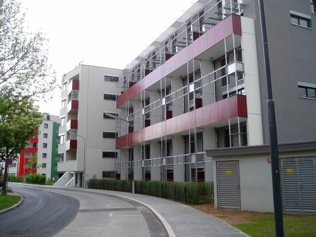 Lange Wohnfassade eines Neubaus mit roten Balkonen entlang einer Straße und Grünstreifen.