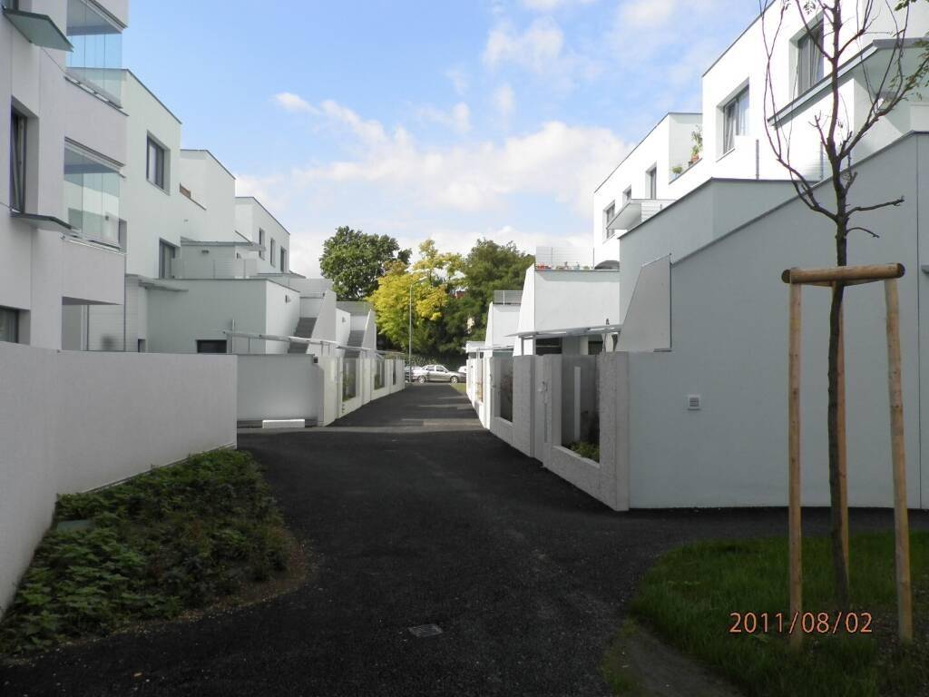 Blick entlang eines dunklen Weges zwischen modernen weißen Wohngebäuden mit viel Grün im Hintergrund.
