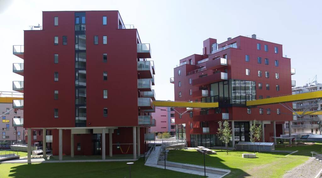 Moderne rote Wohngebäude mit Balkonen und Grünflächen, verbunden durch eine gelbe Brücke.