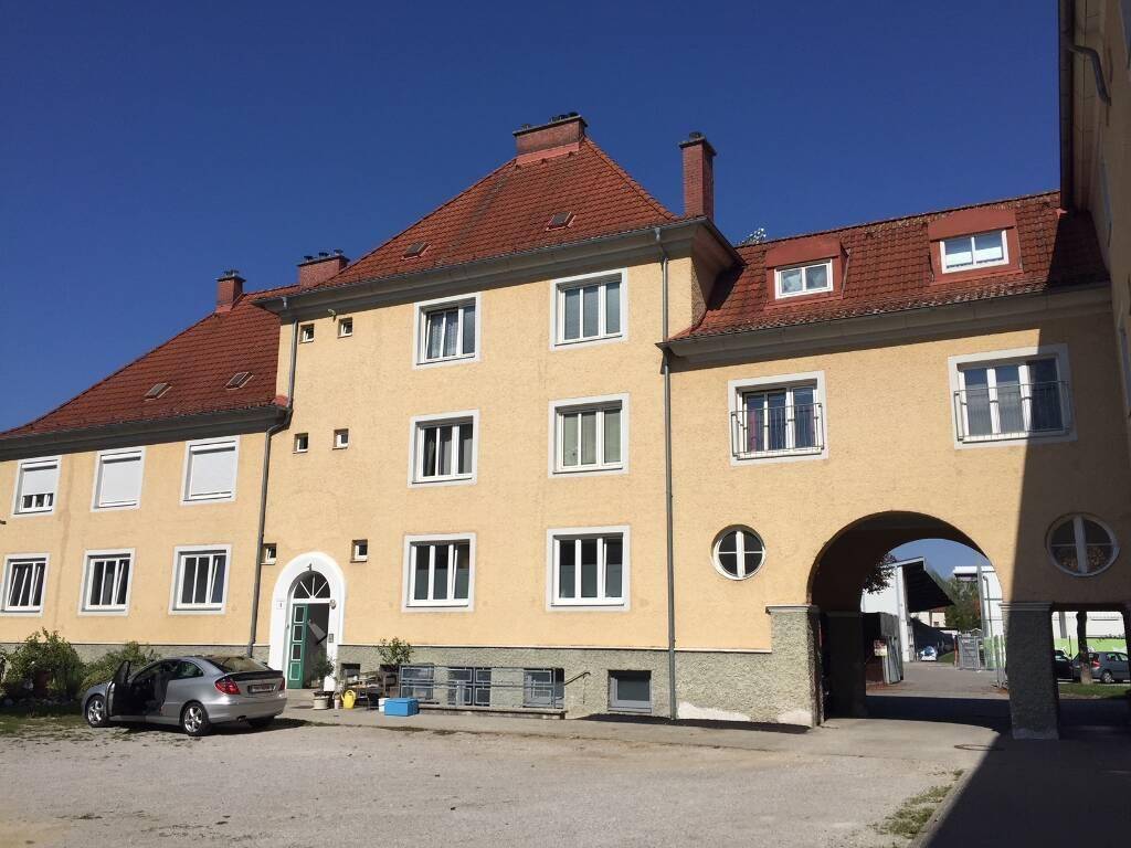Gelbes Mehrfamilienhaus mit rotem Ziegeldach, weißen Fenstern und einem Torbogen, der zu einem Innenhof führt.