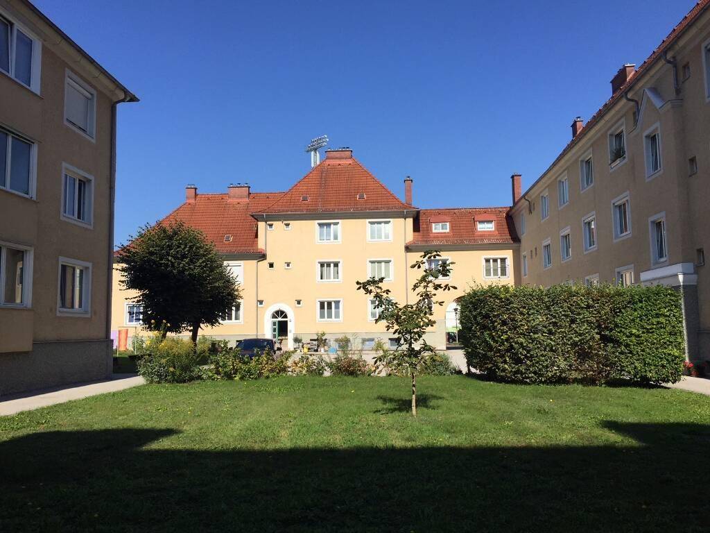 Mehrfamilienhaus mit rotem Ziegeldach, umgeben von einem gepflegten Rasen und hohen Hecken in einem sonnigen Innenhof.