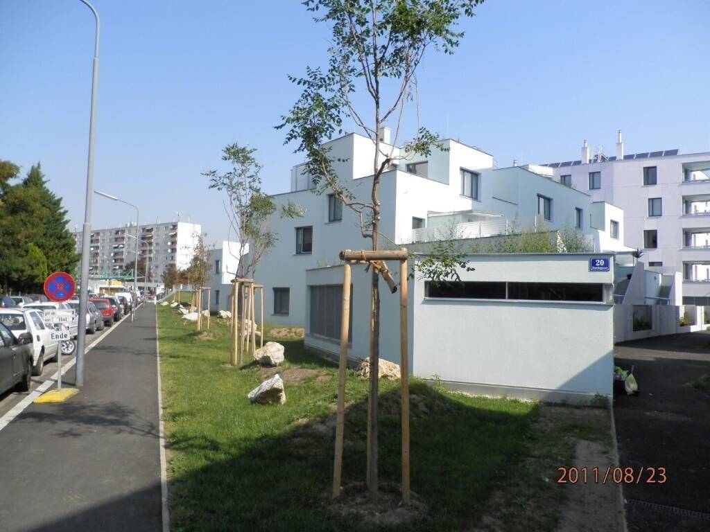 Straßenansicht eines modernen Wohnviertels mit weißen Gebäuden, Grünstreifen und geparkten Autos.