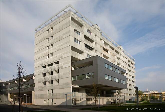 Hohes, modernes Wohngebäude mit markanter Betonfassade und einem auskragenden dunklen Gebäudeteil.
