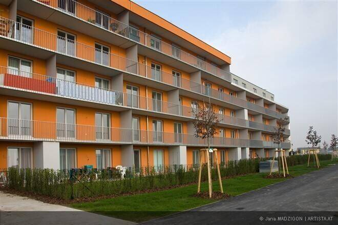 Modernes Wohngebäude mit leuchtend orangefarbener Fassade, zahlreichen Balkonen und gepflegter Grünfläche davor.