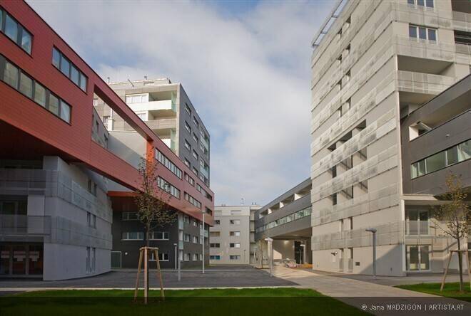 Architektonisch anspruchsvoller Wohnkomplex mit verschiedenen Gebäuden in Beton, Rot und Grau um einen Innenhof.