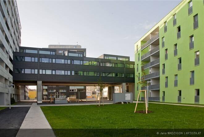 Moderner Wohnkomplex mit einem Gebäude in leuchtendem Grün und einer dunklen Verbindungsbrücke über eine Rasenfläche.