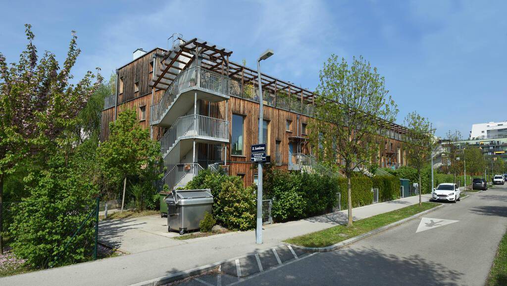 Eckgebäude mit Holzfassade, Außentreppe zu den oberen Etagen und begrünten Dächern in einer Wohnstraße.
