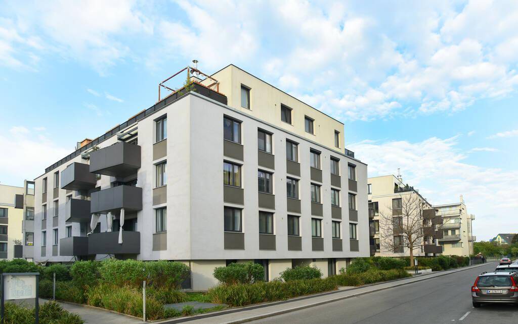 Zeitgenössisches Apartmentgebäude mit weißer Fassade, dunklen Balkonen und gepflegter Außenanlage unter blauem Himmel.