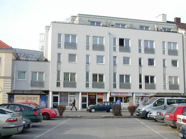 Modernes Mehrfamilienhaus mit Geschäften im Erdgeschoss und Parkplätzen davor in einer städtischen Umgebung.