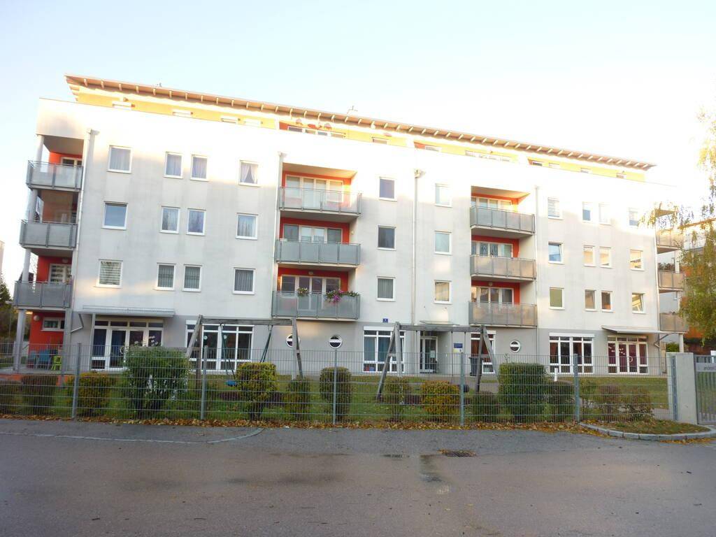 Modernes Mehrfamilienhaus mit hellgrauer Fassade, roten Akzenten und Balkonen sowie einem Spielplatz im Vorgarten.