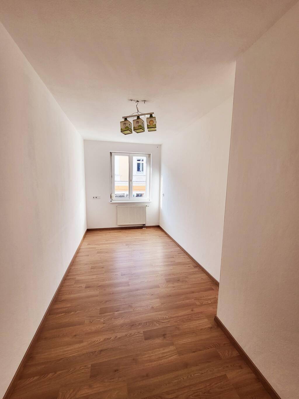 Leeres Zimmer mit Laminatboden und einem Fenster, das viel Tageslicht hereinlässt.