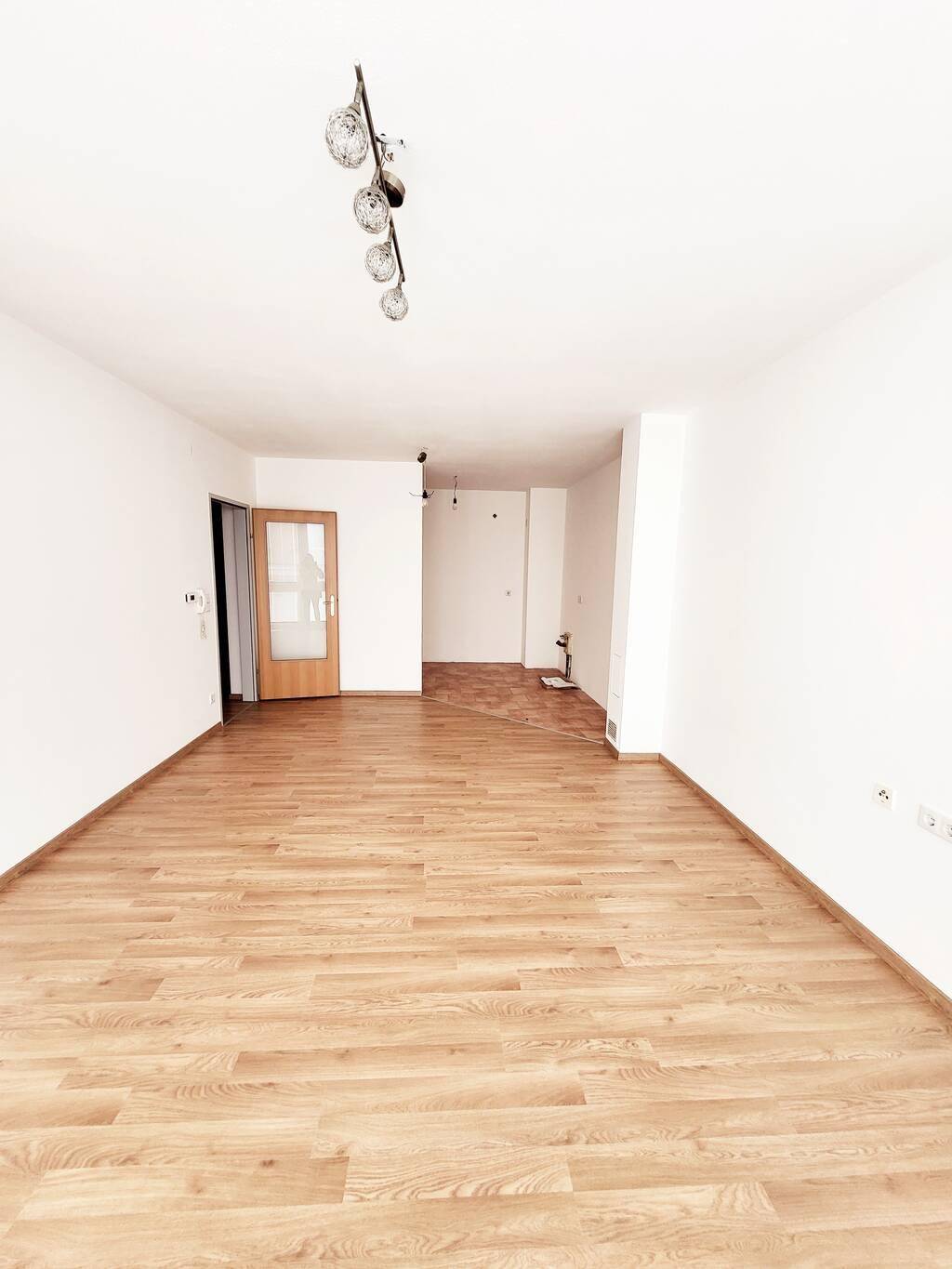 Heller, leerer Raum mit Laminatboden und einer Tür, die in einen weiteren Bereich führt.