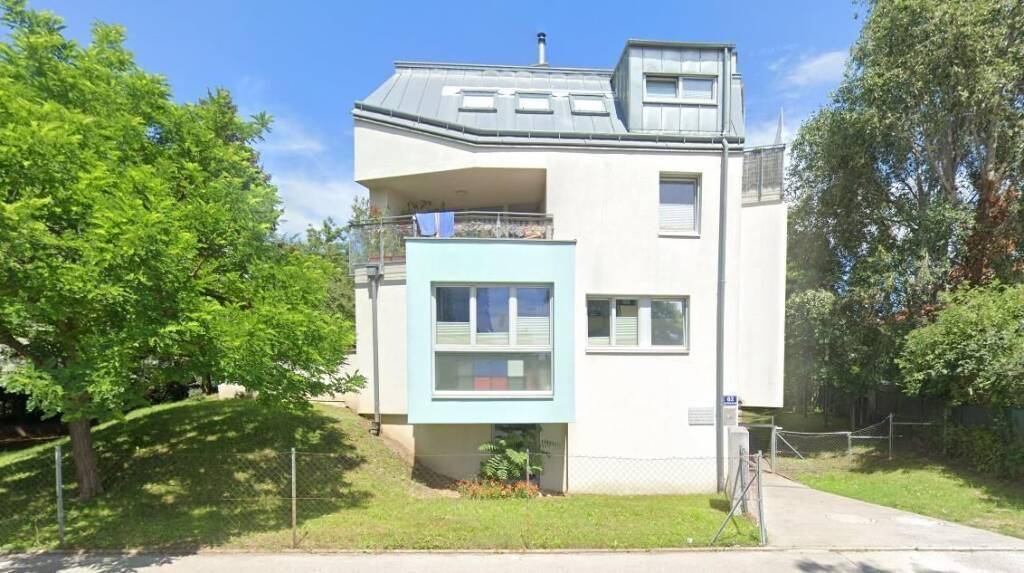 Modernes Wohnhaus mit Mansarddach, Balkon und hellblauer Fassadenakzentuierung, umgeben von Grün.