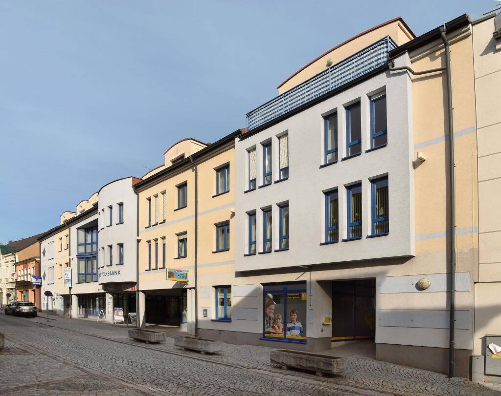 Reihe moderner Gebäude mit hellen Fassaden und blauen Fensterrahmen entlang einer gepflasterten Straße.