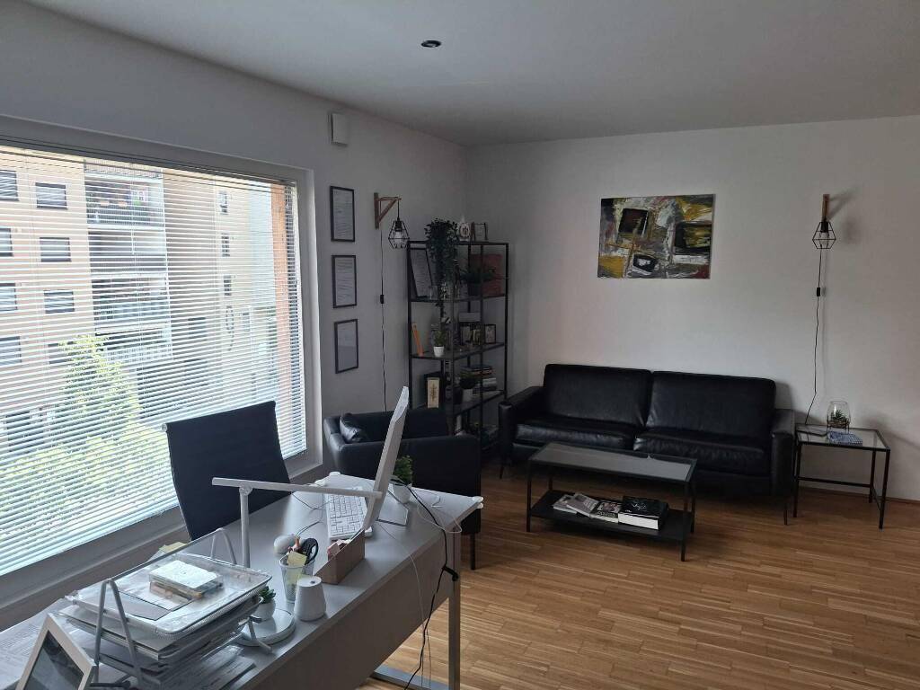 Heller Wohnbereich, der auch als Büro dient, mit Schreibtisch am Fenster, Ledersofa und Holzfußboden.