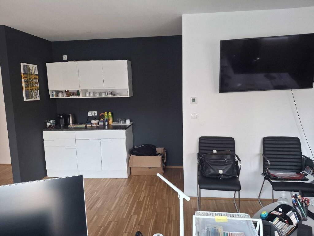 Kleine Küchenzeile mit weißen Schränken an dunkler Wand, angrenzend an einen Wohnbereich mit Fernseher.