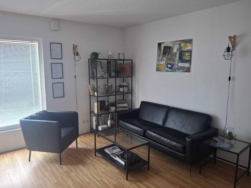 Gemütliches Wohnzimmer mit schwarzem Ledersofa, Sessel, Glas-Couchtisch und Holzfußboden in heller Umgebung.