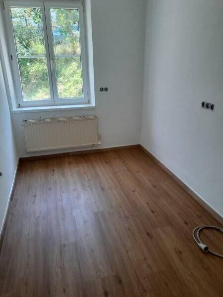 Helles Zimmer mit Laminatboden, weißen Wänden und Fenster mit Ausblick ins Grüne.