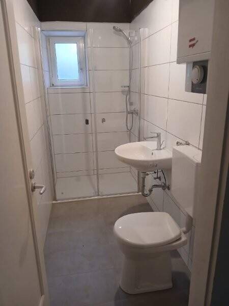 Helles Badezimmer mit Duschkabine, WC, Waschbecken und Fenster, ausgestattet mit weißen Fliesen.