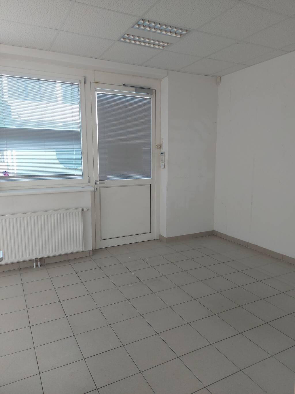 Leerer Büroraum mit Fliesenboden, Fenster mit Jalousien und einer Tür, die nach außen führt.