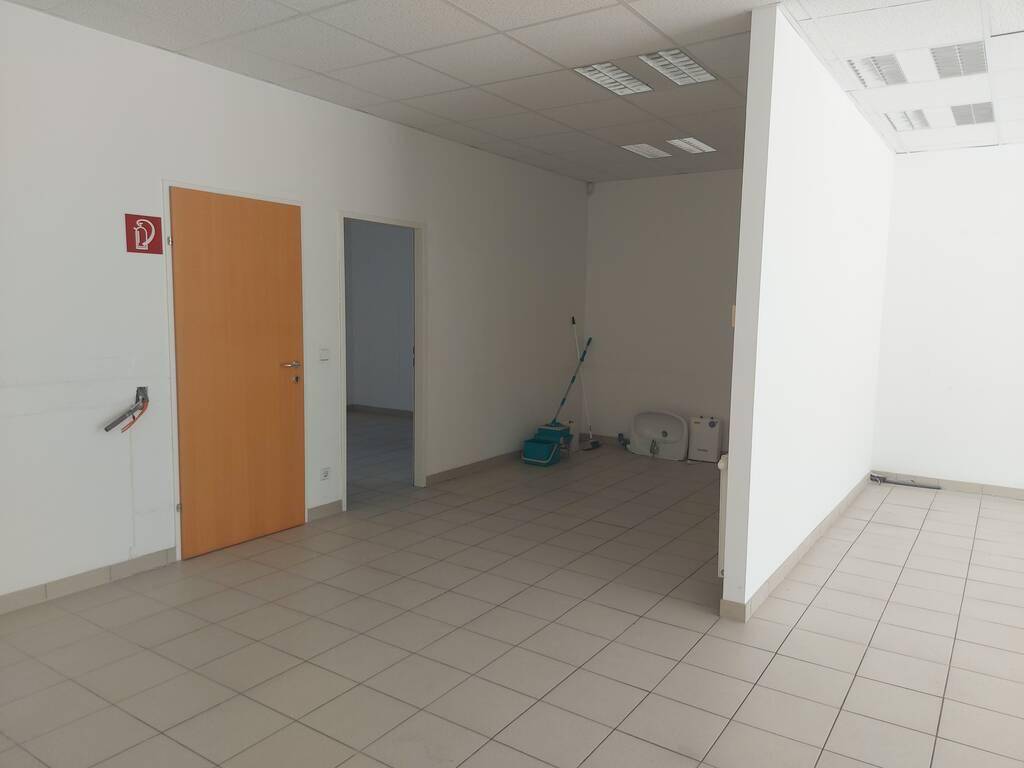 Großer, leerer Geschäftsraum mit Fliesenboden, einer Holztür und einem offenen Durchgang zu einem weiteren Bereich.