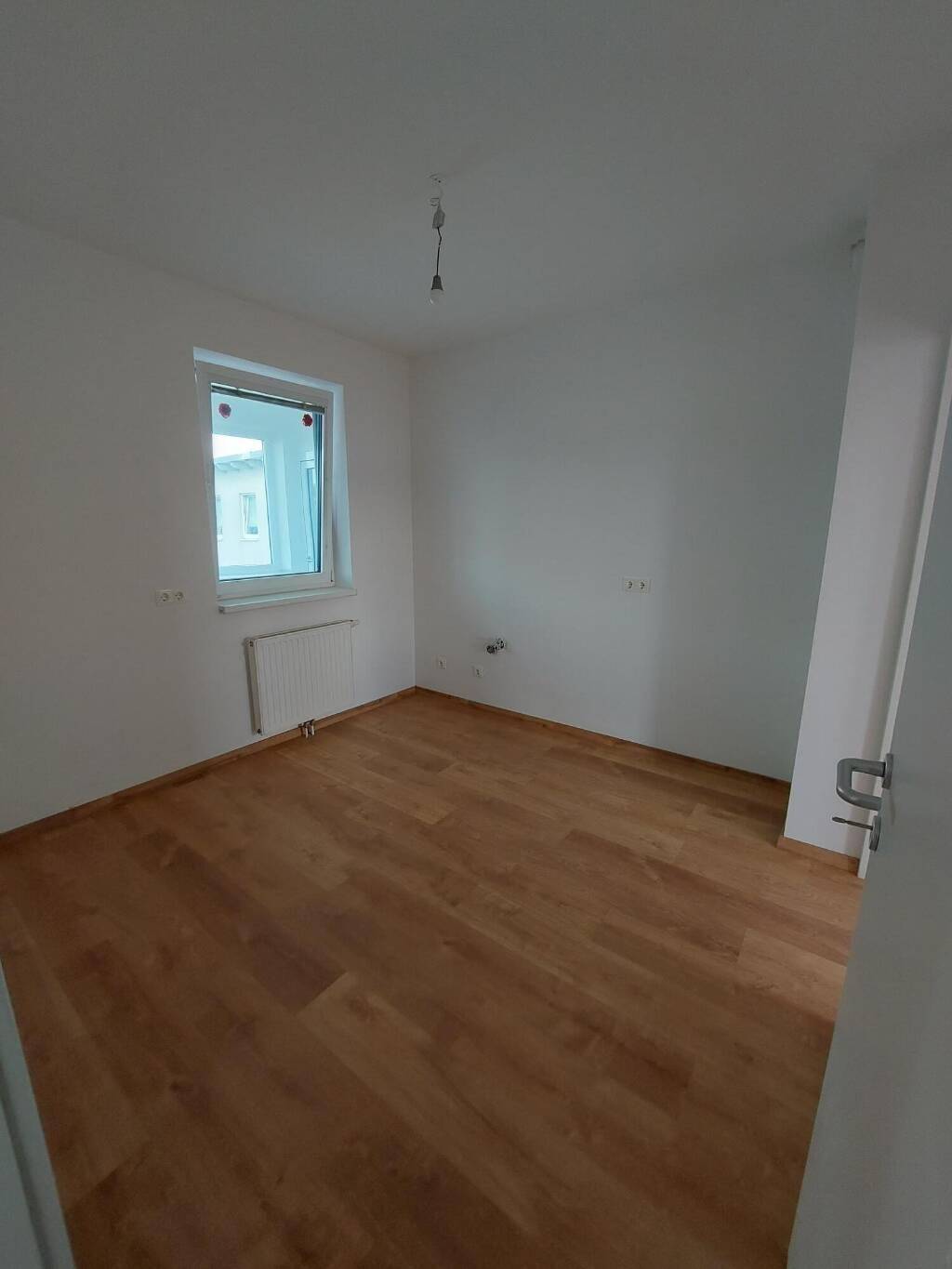 Helles, leeres Zimmer mit Laminatboden, weißen Wänden und einem Fenster, ideal als Schlaf- oder Arbeitszimmer.