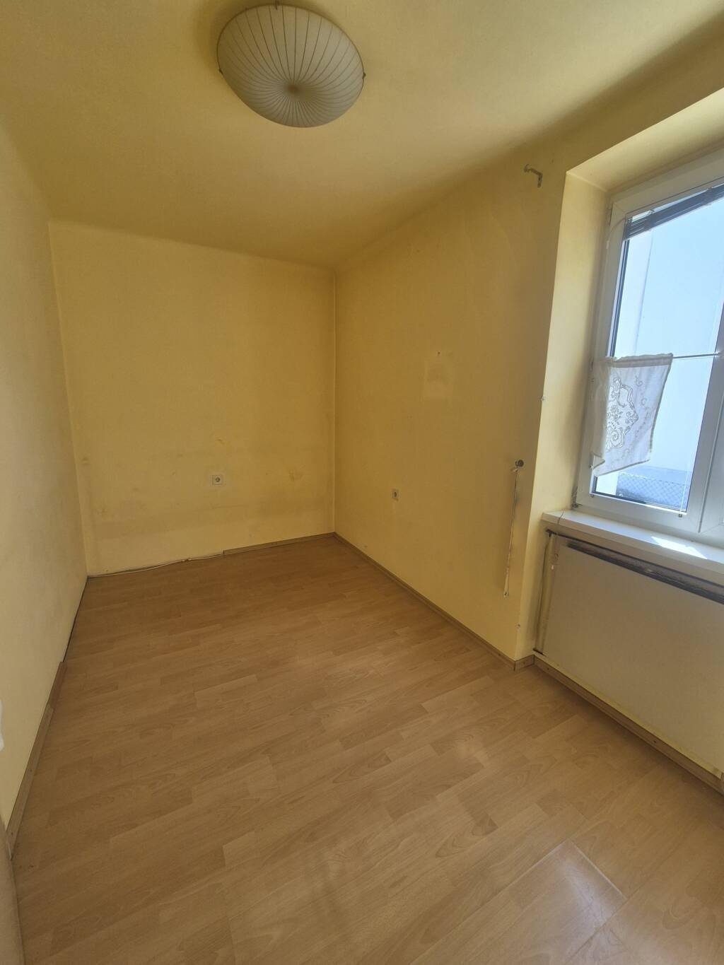 Leeres Zimmer mit hellem Laminatboden, gelben Wänden und einem Fenster mit Heizkörper darunter.