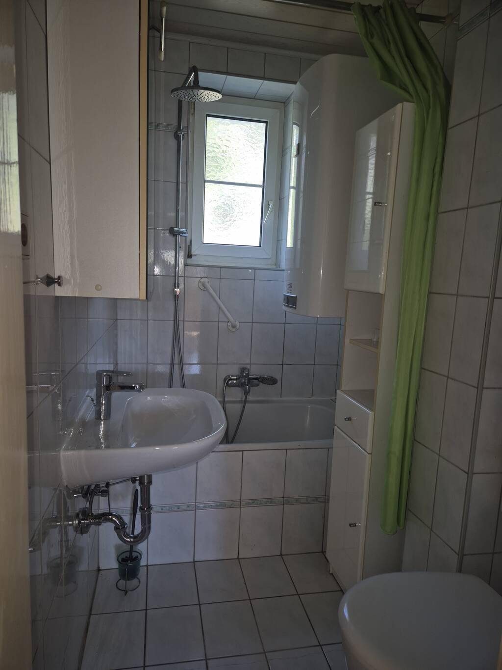 Kleines Badezimmer mit weißen Fliesen, Badewanne, Dusche und Fenster für Tageslicht.