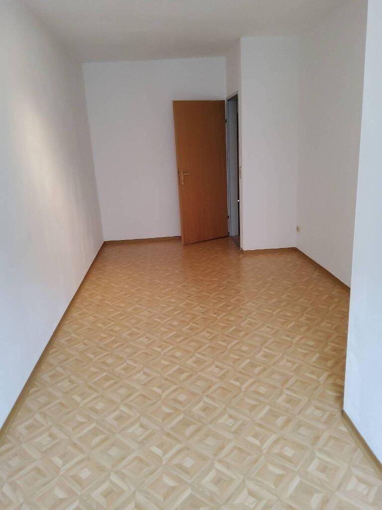 Helles, leeres Zimmer mit Parkettboden und einer geöffneten Holztür.