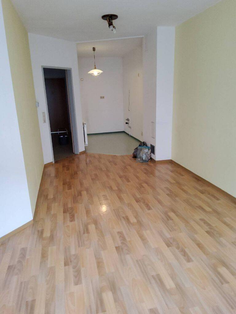 Geräumiger, heller Raum mit glänzendem Parkettboden und Zugang zu einem Flurbereich.