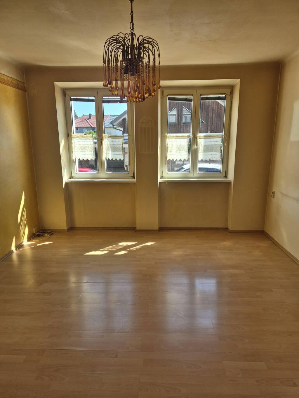 Helles Wohnzimmer mit Laminatboden, zwei Fenstern und einem dekorativen Kronleuchter.