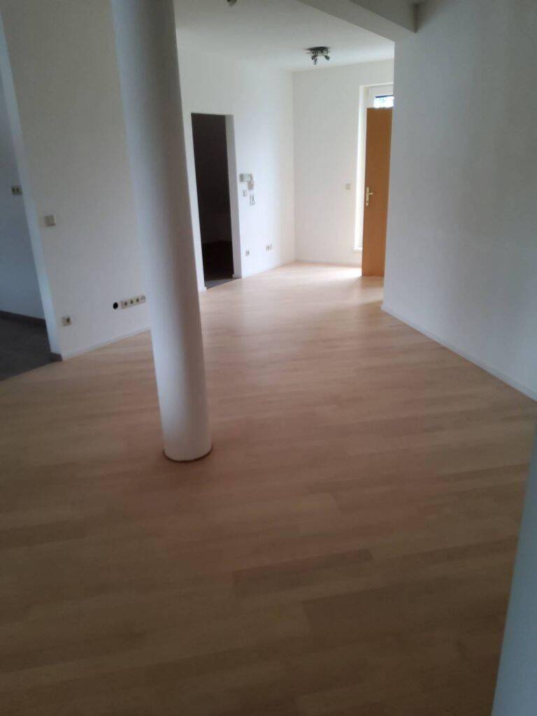 Heller, leerer Raum mit hellem Holzboden und einer tragenden Säule in der Mitte.