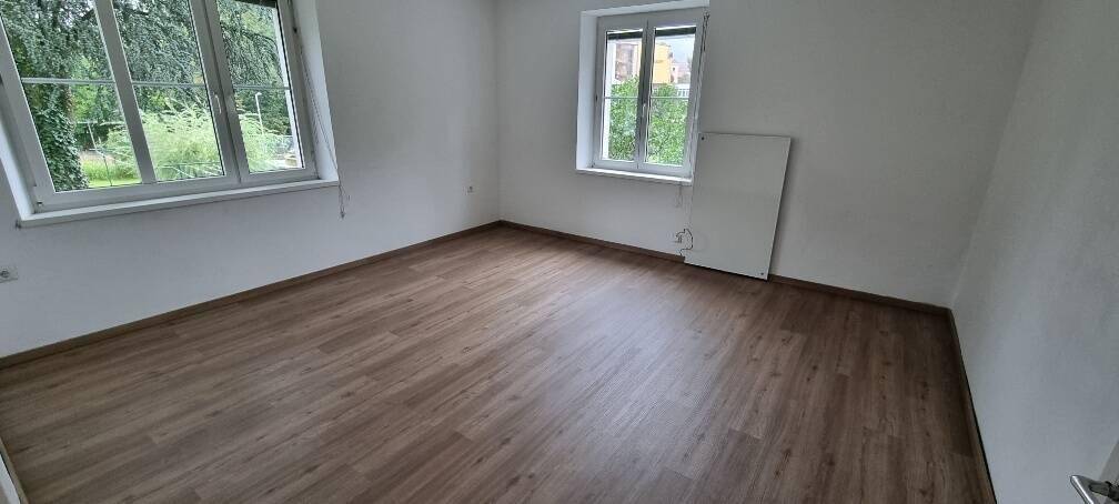 Geräumiger leerer Raum mit hellem Laminatboden und zwei großen Fenstern mit Blick ins Grüne.