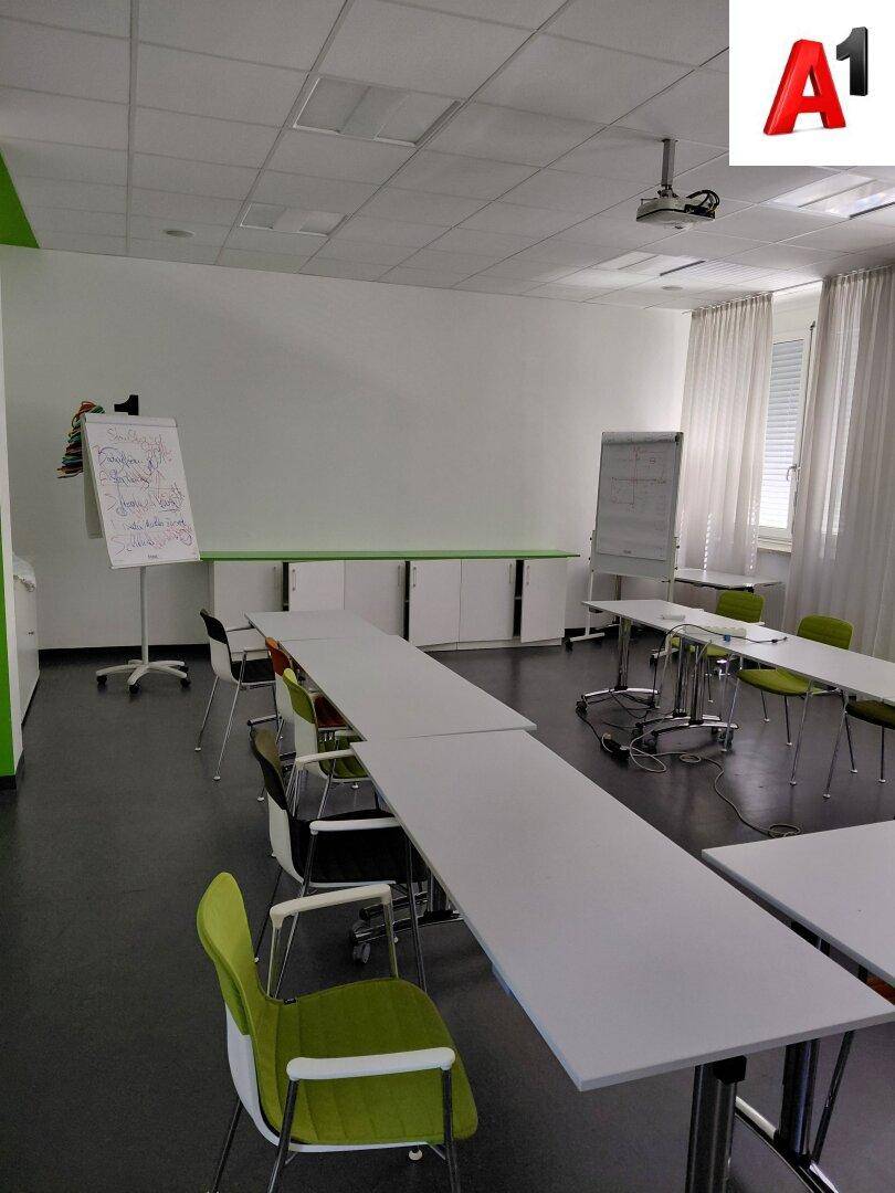 Großer Konferenzraum mit langen Tischen, grünen Stühlen und Whiteboards für Präsentationen.